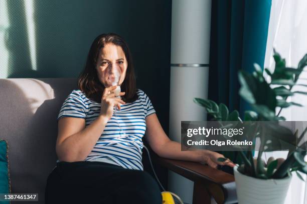 brown haired woman using nebulizer for inhalation, looking at camera, breathing problems. sitting indoors near light window - atemhilfe stock-fotos und bilder