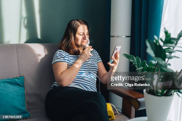 brown haired woman using nebulizer for inhalation, looking for information in smartphone. sitting in living room near light window - respiratory disease stock pictures, royalty-free photos & images