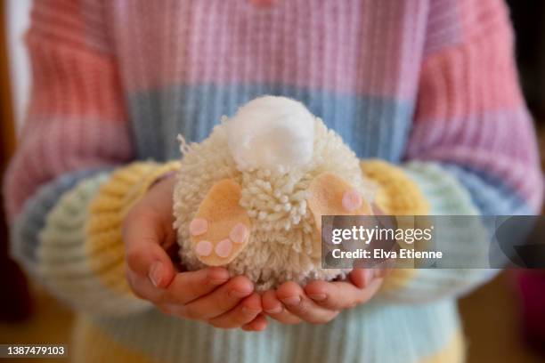 close up of a homemade easter bunny themed pom-pom held in the hands of a child wearing a multicoloured knitted sweater. - pom pom stock-fotos und bilder