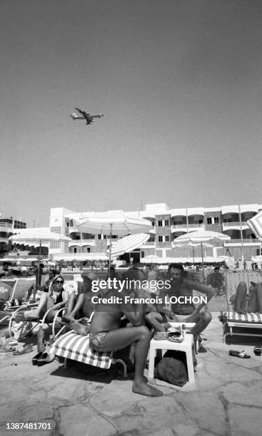 Touristes sur des transats près d'une piscine dans un complexe hôtelier et avion de ligne dans le ciel en juillet 1978 à Beyrouth.