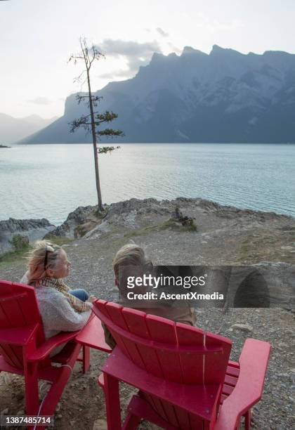 mature couple relax in red arm chairs on lakeshore - lake minnewanka stockfoto's en -beelden