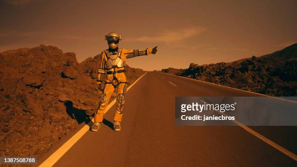 astronaut looking for a lift on mars. hitchhiking on a mountain road. surreal scene - ironia imagens e fotografias de stock