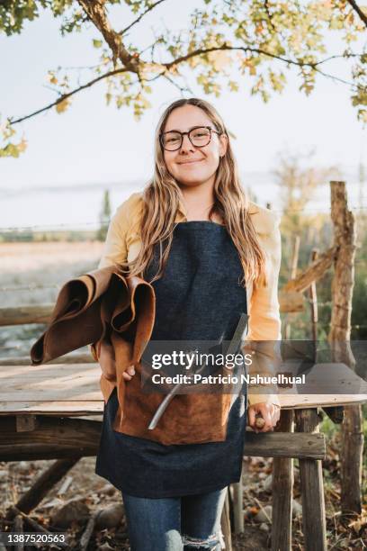 young artisan craftswoman holding leather sheets on her arm and carrying different tools in her denim apron pocket. independent small business. workers' day. female artisan - 1 de maio - fotografias e filmes do acervo