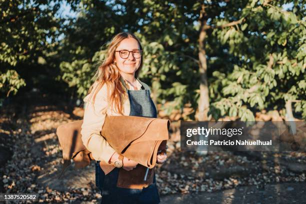 young artisan craftswoman in a tree plantation,  holding leather sheets on her arm and carrying different tools in her denim apron pocket. independent small business. workers' day. female artisan - chilean ethnicity stock pictures, royalty-free photos & images