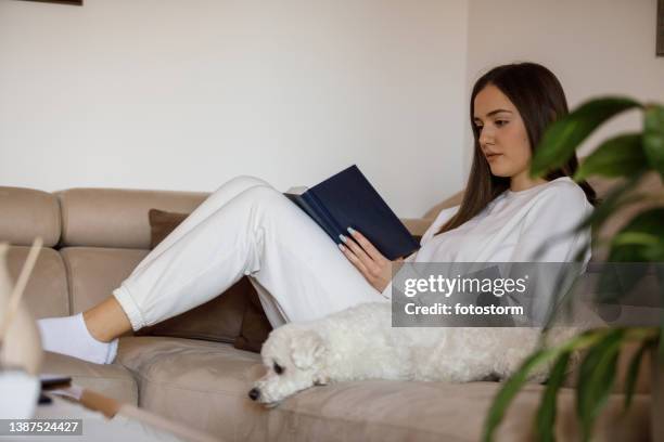 ソファでくつろぎ、本を読むのを楽しんでいる若い女性 - トレーニングウェア ストックフォトと画像