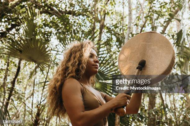 una donna multirazziale che suona il tamburo sciamanico nella foresta pluviale contro la luce del sole - solfato di cobalto foto e immagini stock