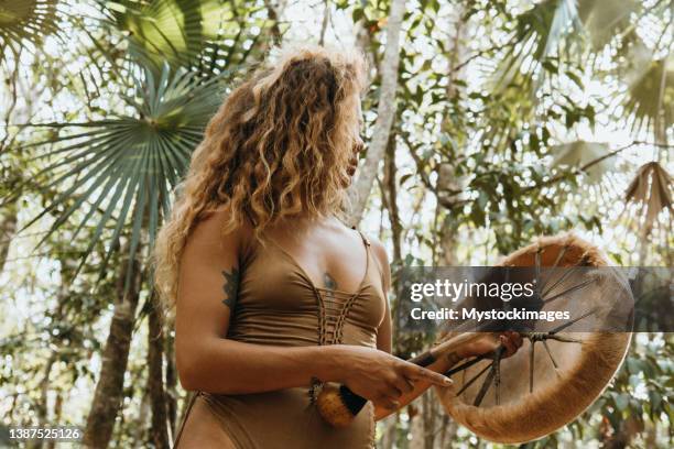 una donna multirazziale che suona il tamburo sciamanico nella foresta pluviale contro la luce del sole - solfato di cobalto foto e immagini stock