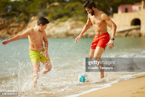 a father and son play by the water's edge with a soccer ball. - swimming shorts stock pictures, royalty-free photos & images