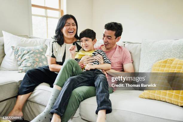 medium wide shot of smiling and laughing parents sitting on couch in living room with son on lap - mom sits on sons lap stock-fotos und bilder