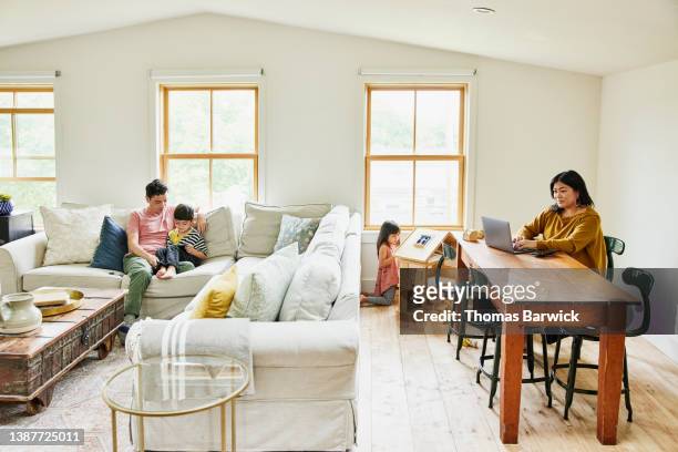 wide shot of mother working on laptop while family plays in living room - living room young couple stock-fotos und bilder