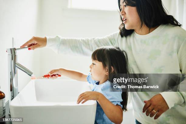 Medium shot of mother helping daughter put water on toothbrush in bathroom