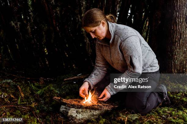 a woman stokes a small fire she has lit in front of a survival shelter on a rainy day - stoking stock pictures, royalty-free photos & images