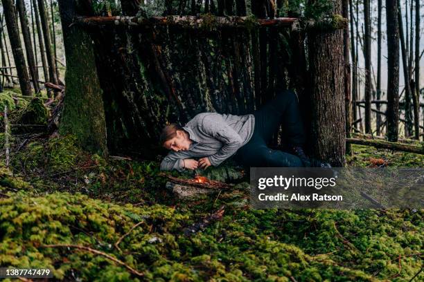 a woman laying in a makeshift survival shelter blows on the flame of her small campfire - makeshift shelter stock pictures, royalty-free photos & images