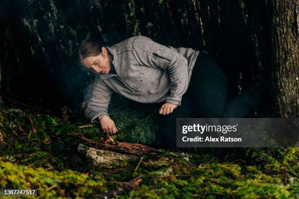 a woman lays inside of a makeshift survival shelter beside a small fire on a rainy day - makeshift shelter stock pictures, royalty-free photos & images