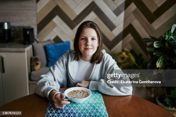 a 12-year-old girl with brown hair sits at home at the table and has breakfast muesli with milk - homegirl stock-fotos und bilder