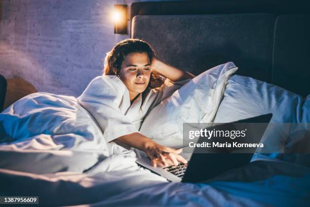 young woman laying in bed and using laptop at the night. - work video call stock pictures, royalty-free photos & images