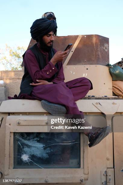 Young Talib in black turban and wearing sandals holds a smartphone in one hand, browses the social media while sat comfortably next to the shattered...