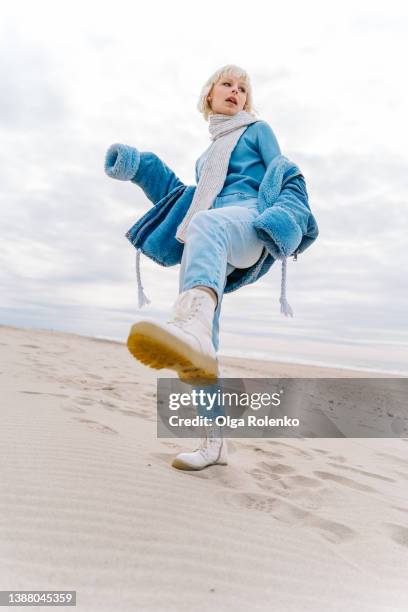 young modern blond woman in warm jeans outfit and a scarf posing on a sandy beach. sea background - hipster girl imagens e fotografias de stock