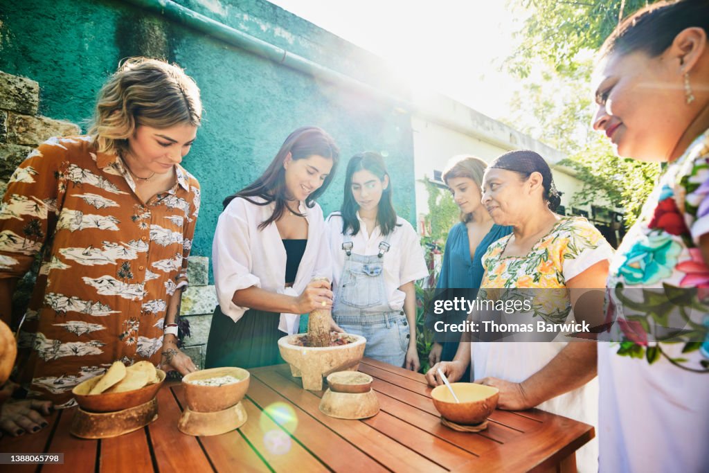 Medium wide shot of smiling friends grinding salsa in mortar during traditional Mayan cooking class