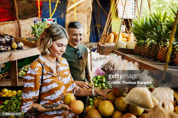 medium shot of smiling couple picking out fruit at local market while on vacation - tourist market stockfoto's en -beelden