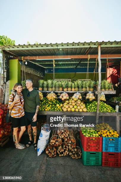 Medium shot portrait  of smiling embracing couple standing in front of fruit stand while on vacation