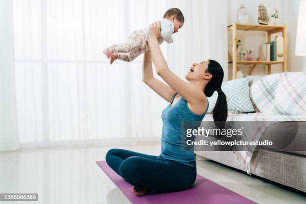side view asian female parent is sitting on the floor and raising her daughter up high. cheerful woman interacting with cute infant at home. real moments - full height stock pictures, royalty-free photos & images