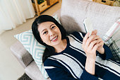 pretty lady reclining on sofa with phone in hand is smiling at camera. portrait asian young female is relaxing by phubbing at home. technology and lifestyle.