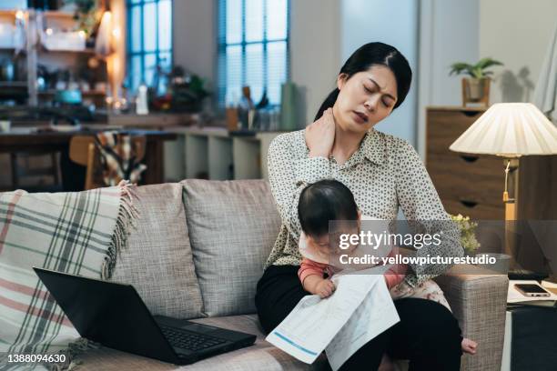 retrato de la madre trabajadora asiática que sostiene al bebé tiene tensión en el cuello. mamá milenaria torciéndose el cuello mientras cuidaba a su hijo. encantadora niña está mirando el documento en la mano de su madre - massage room fotografías e imágenes de stock