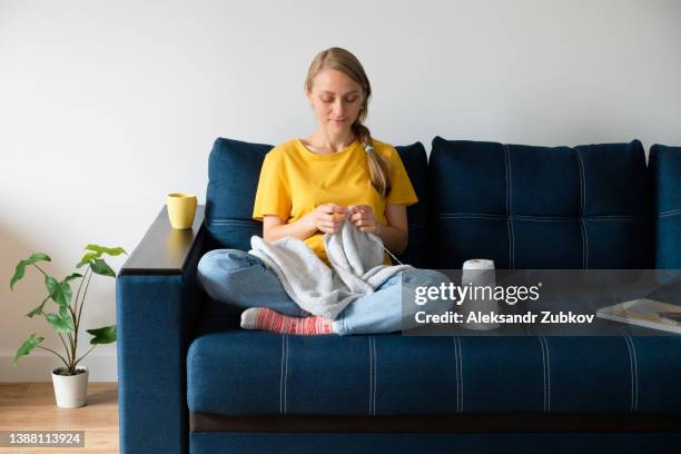 the girl is holding a product and knitting needles in her hands. there is a mug of coffee or tea next to it. a woman knits a woolen sweater or scarf, sitting on the couch at home. the concept of hobbies, creativity, needlework and manual work. copy space. - knitting needles stock-fotos und bilder