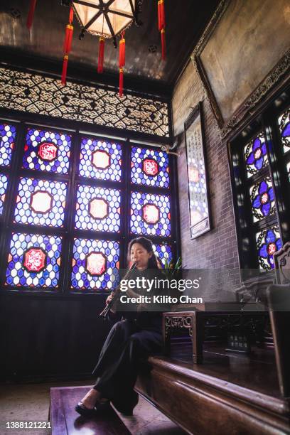 senior asian woman playing shakuhachi flute in a traditional chinese house - chinese window pattern stockfoto's en -beelden