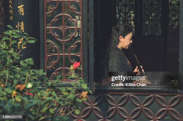 senior asian woman practicing shakuhachi flute by the window of a traditional chinese house - chinese window pattern stockfoto's en -beelden