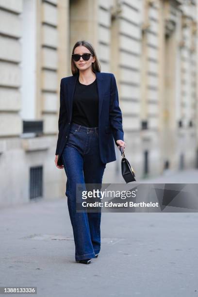 Diane Batoukina wears black sunglasses, a black t-shirt, a navy blue blazer jacket from Saint Laurent Paris, a black shiny leather with gold V logo...
