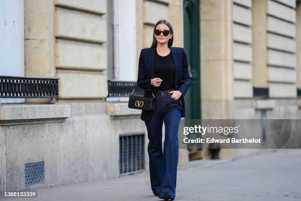 Diane Batoukina wears black sunglasses, a black t-shirt, a navy blue blazer jacket from Saint Laurent Paris, a black shiny leather with gold V logo...