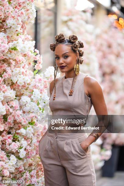 Alicia Aylies wears a gold large face print pattern pendant earring, gold rings in the hair, a brown cropped tank-top, matching high waist brown wide...