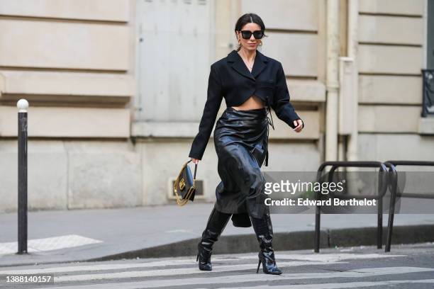Katie Giorgadze wears gold earrings, black sunglasses, a gold chain pendant necklace, a black cropped blazer jacket, a black shiny leather high waist...