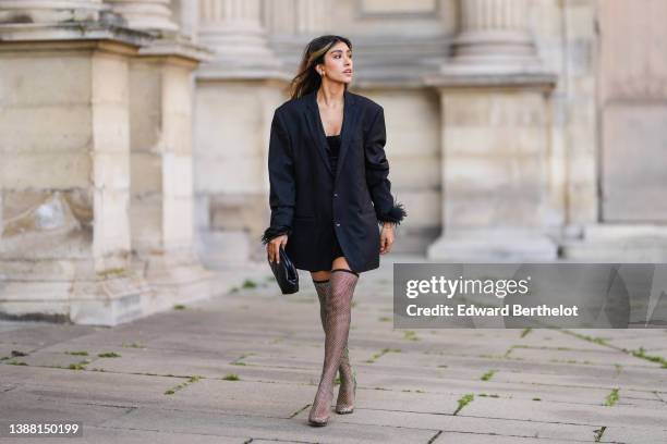 Angela Gonzalez wears gold earrings, a black oversized blazer jacket with feather wrists, a black shiny leather quilted clutch, black cyclist shorts,...