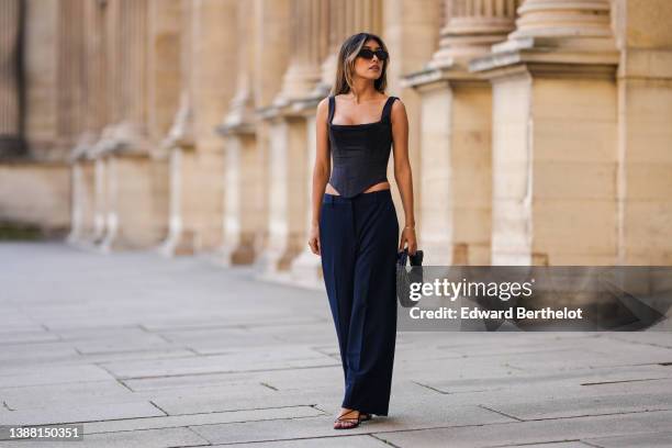 Angela Gonzalez wears a black long blazer jacket, black sunglasses, gold earrings, a black square-neck / corset tank-top, navy blue large suit pants,...