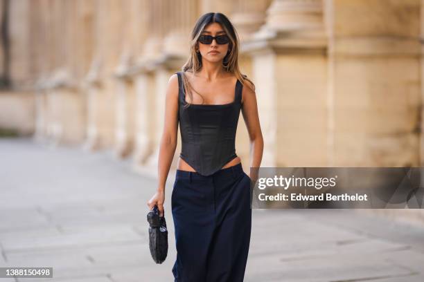 Angela Gonzalez wears a black long blazer jacket, black sunglasses, gold earrings, a black square-neck / corset tank-top, navy blue large suit pants,...