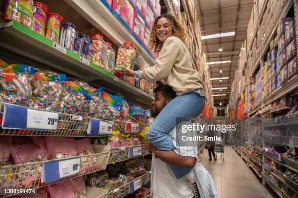 young woman on man's shoulders shopping for candy - couple in supermarket stock-fotos und bilder