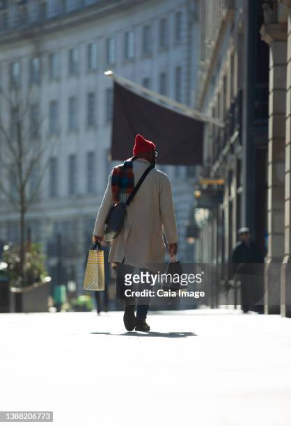 man with shopping bags walking on sunny city sidewalk - trench coat back stock pictures, royalty-free photos & images
