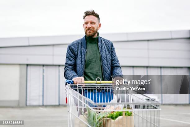 smiling young man with a shopping cart in front of a big shop .consumerism / shopping concept. - shopping cart stock pictures, royalty-free photos & images