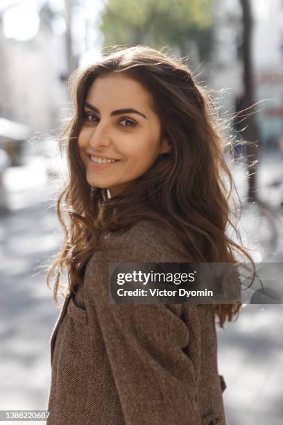 cute girl with brown eyes in a brown coat turns to look at the camera and smiling. - cabello castaño fotografías e imágenes de stock