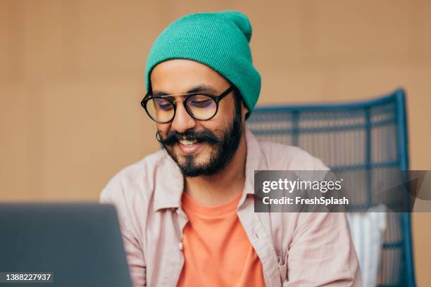 a happy casually dressed man with moustaches sitting in his office at computer and chatting - hipster developer stockfoto's en -beelden