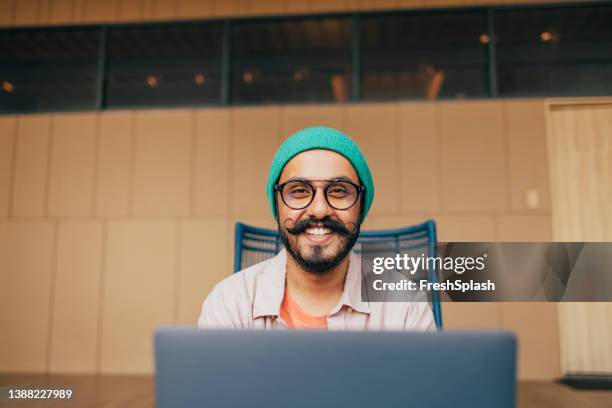 a portrait of a happy casually dressed man with moustaches looking at camera while sitting in his office at computer - bruine hoed stockfoto's en -beelden