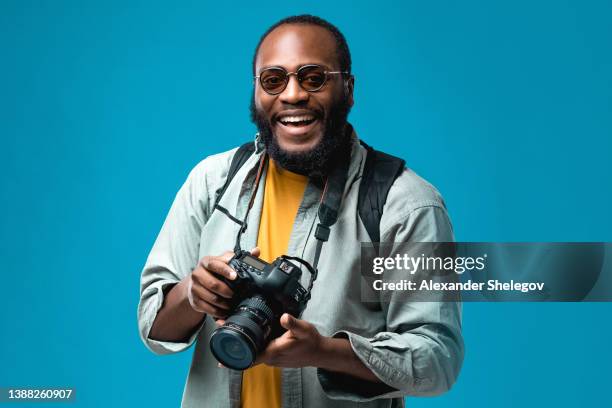 portrait african-american man at the studio with colored background. black person wearing sunglasses and blue t-shirt. traveling and tourism concept. photographer with professional digital camera - photographer stock pictures, royalty-free photos & images