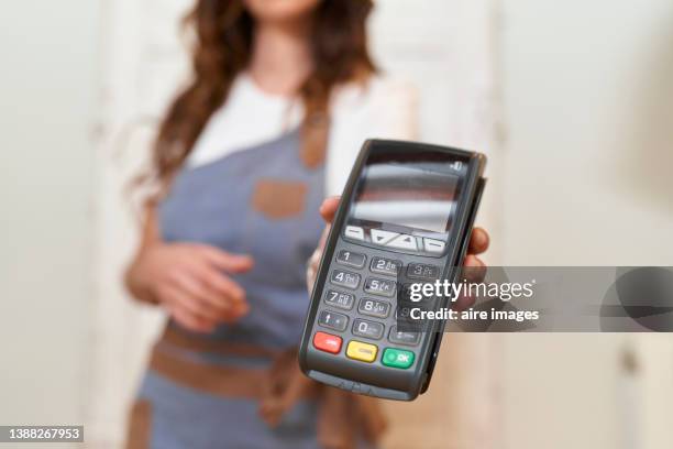 close-up of hand of an unrecognizable woman with apron standing in the kitchen of her house holding payment terminal to enter payment for her purchase with a credit card - verkehrsgebäude stock-fotos und bilder