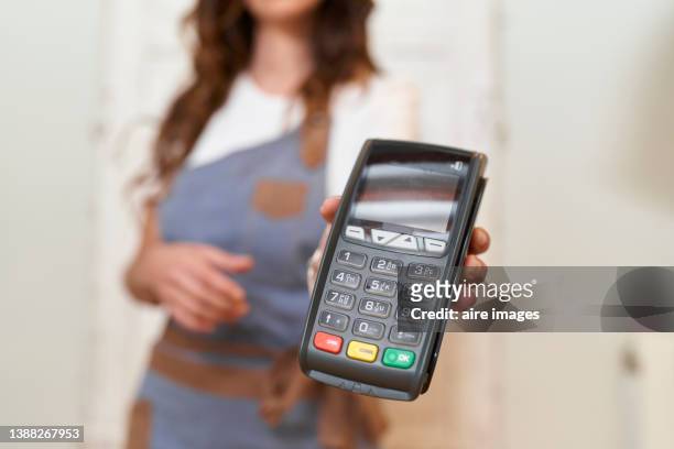 close-up of hand of an unrecognizable woman with apron standing in the kitchen of her house holding payment terminal to enter payment for her purchase with a credit card - credit card terminal stock pictures, royalty-free photos & images
