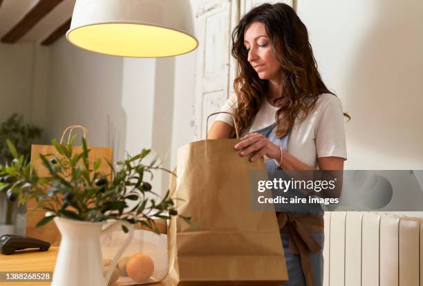 front view of side view of a young caucasian woman in casual clothing coming home with grocery bags to unpack the groceries on her kitchen table at home - sachet en papier photos et images de collection