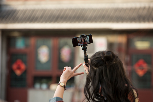 Back view of long curly hair young Chinese lady taking selfies with selfie stick - stock photo