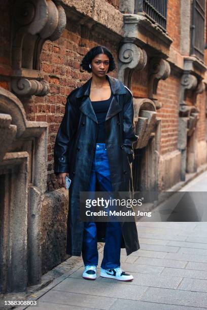 British Jamaican-Sri Lankan model Shivaruby Premkanthan wears Apple Airpods, a long black leather Chillie London Vintage trench, black top, blue...